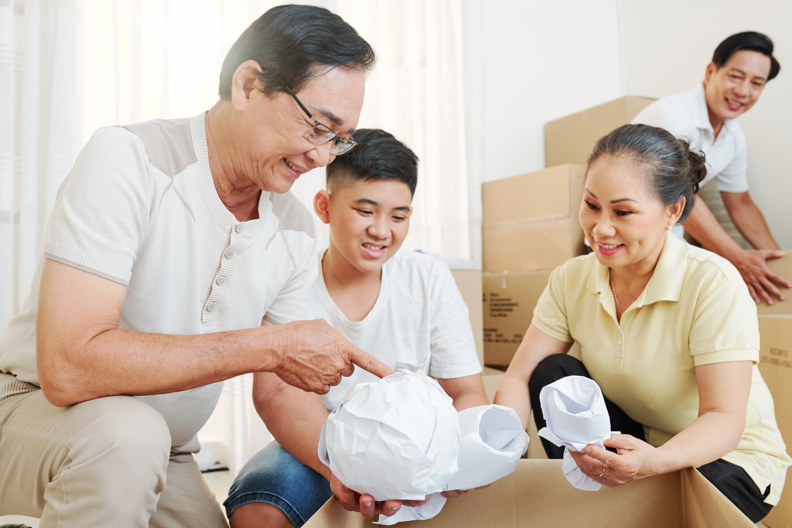 Cheerful grandparents explaining to boy how to wrap fragile and significant things before packing the in cardboard boxes.